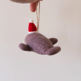 A hand holds a felt manatee ornament, suspended by a jute string against a plain, light background. The charming purple felt manatee ornament features a festive red and white Santa hat, adding a touch of gentle whimsy.