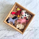 A playful wool felt Milk Cow ornament, white and black with a pink nose, rests in a square cardboard box filled with shredded paper on a bright marble surface. This handmade felt ornament, crafted from 100% natural New Zealand wool, evokes rustic charm and playful creativity.