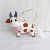 An ethically hand-made wool felt Milk Cow ornament is displayed against a clean white background, suspended by a jute string, ready for a Christmas tree. This whimsical ornament features a white body with evenly spaced brown spots, a pink muzzle, brown hooves, and grey horns.