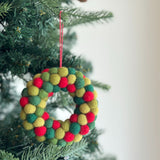 A wool felt mini pompom heart wreath ornament hangs festively from a pine Christmas tree. This cheerful, handcrafted Christmas tree ornament features tightly packed red, green, and yellow pompoms forming its heart shape, complete with a red string for hanging. It evokes warmth and holiday cheer.