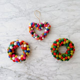 A cheerful wool felt Mini Pompom Heart Wreath ornament rests on a mottled white and gray marble background. This petite heart-shaped ornament features a jute string hanger and is vibrantly adorned with red, white, pink, green, and blue pompoms.