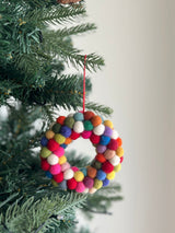 A vibrant Wool Felt Mini Pompom Wreath Christmas Ornament hangs from a green Christmas tree branch, suspended by a thin red string. This petite, heart-shaped felt ornament is adorned with tightly packed mini pompoms in red, pink, white, blue, green, orange, and yellow, creating a festive and cheerful look.
