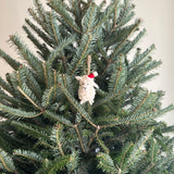 A handmade wool felt mini sheep ornament hangs on a festive green Christmas tree. This cream-colored sheep features a fluffy body, a stitched black smiling mouth, and a red and white Santa hat jauntily placed on its head. It adds a whimsical touch to the holiday decor.