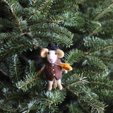 A handmade felt mouse boy ornament hangs on a fresh green Christmas tree. This needle felted wool mouse features a white face, tan ears, and a pink stitched nose. Dressed in a brown vest with buttons and a black hat, it playfully holds a yellow leaf. A black string is attached to the hat for easy hanging.