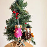 A charming needle felted mouse ornament, suspended by a white string, hangs on a small Christmas tree adorned with bright red berries. The tree, resting on a round wooden table, has a rustic burlap-wrapped base. The 3D mouse ornament features a brown coat with white polka dots, complemented by a scarf and hat, adding a whimsical touch to the festive display.