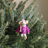 A charming needle felted mouse Christmas ornament, featuring a pink scarf and sweater, hangs from a vibrant green Christmas tree branch. The handcrafted felt mouse has a light beige face and fluffy ears, and is dressed in a cheerful pink knit hat and matching scarf. Its pink sweater is adorned with a white snowflake design, and a white string allows it to hang.