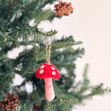 A cheerful felt mushroom ornament hangs on a green Christmas tree adorned with green needles and pine cones. The felt mushroom ornament features a bright red cap with white dots and a pale pink stem, with a jute string attached for hanging.
