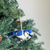 A needle felted blue jay ornament hangs gracefully on a green pine tree branch, evoking a natural and joyful atmosphere. This felt blue jay is predominantly blue with white accents on its face and underbelly, a black beak, and delicate felted details forming its legs. A jute string is attached for hanging.