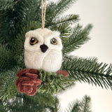 This charming needle felted owl ornament is displayed hanging on a Christmas tree. The white owl has a brown beak and prominent yellow eyes with black pupils, perched on a green and brown cypress tree. A jute string is visible at the top for easy hanging.