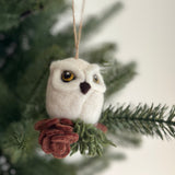 A handcrafted needle felted owl ornament is featured against a soft, out-of-focus backdrop of green Christmas tree branches, creating a festive scene. This charming white owl ornament has a fluffy, textured body with a small black beak and yellow eyes. It is perched on a wood-themed base with green foliage. A jute string at the top suggests it is ready to be hung as a Christmas tree ornament.