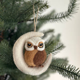 A handcrafted needle felted owl ornament hangs gracefully on a lush, green Christmas tree. The charming brown owl, made from sections of white, light brown, and dark brown felt, is perched on a white crescent moon. Its black-accented eyes are visible, and a jute string is attached for easy hanging.