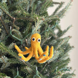 A handcrafted yellow octopus felt ornament hangs on a Christmas tree adorned with delicate lights. This vibrant yellow octopus features eight legs detailed with small blue dots and big white eyes with black pupils. A yellow cotton string is attached to its head, allowing it to hang gracefully, adding a cheerful holiday touch.
