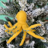 A yellow octopus felt ornament hangs joyfully on a snow-flocked Christmas tree. This whimsical wool felt octopus ornament features a rounded head and eight posable wire legs. A matching yellow cotton string allows it to blend seamlessly with the festive branches and pine cones.