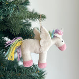 A pastel unicorn felt ornament hangs from a green Christmas tree branch by a jute string. This soft, white wool felt unicorn features a pink snout, pink hooves, and soft white felt wings. Its yarn tail and fluffy mane display playful pastel shades of yellow, light blue, and lavender, creating a whimsical and joyful Christmas tree ornament.
