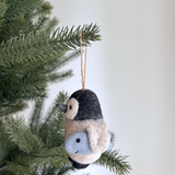 A delightful felt penguin ornament, featuring black and beige tones with a light blue fish detail, hangs from a rustic jute string on a lush green Christmas tree. This handcrafted felt ornament adds a charming, festive touch.