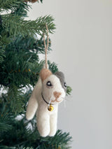 A playful wool felt cat ornament hangs on a Christmas tree. The handmade felt cat ornament is predominantly white with tan and gray patches on its head, featuring intricate hand embroidery for its whiskers and expression. It has a black collar, a small golden bell, and a jute string for easy hanging.