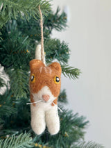 A playful wool felt cat ornament hangs from a vibrant green Christmas tree branch, adding a cozy, festive touch. This soft, textured cat features a brown head with white facial accents, a white body, and charming details like round eyes, black pupils, a pink nose, and delicate white whiskers. A jute string on top allows it to dangle gracefully.