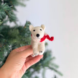 A handmade felt polar bear ornament with a red-and-white scarf is gently held by a hand against a festive Christmas tree. The charming white polar bear features tiny black eyes, a black nose, rounded ears, and detailed paws with embroidered claws. Its red-and-white scarf adds a delightful, festive touch, making it a unique Christmas tree ornament.
