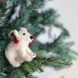 A felt polar bear ornament is perched on a vibrant green pine branch of a lush Christmas tree. This charming polar bear ornament has a rounded white body, black nose, and black feet. A festive red and white braided cotton string, resembling a scarf, is tied around its neck, and a jute string is attached for hanging.