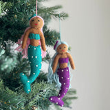 Two vibrant handcrafted felt Pride Mermaid ornaments hang gracefully on a Christmas tree with vivid green needles. One mermaid features light brown skin, pink hair, a turquoise top, and a matching tail adorned with white dots, suspended by a pink string. The second mermaid has light brown skin, light blue hair, a purple top, and a coordinating tail with white dots, hanging from a light blue string. These unique Christmas ornaments embody self-expression and radiant joy.