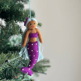 A handcrafted felt Pride Mermaid ornament hangs suspended by a light blue string on a lush, full Christmas tree against a soft light gray backdrop. This vibrant felt Pride Mermaid features a light brown upper body, a bold purple top, and a matching tail adorned with small white dots. Long, vibrant light blue hair flows from beneath its purple and light blue cap, creating a cheerful, whimsical, and joyful Christmas tree ornament.