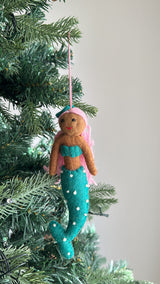 A handcrafted felt mermaid ornament hangs on a Christmas tree, set against a light gray background. This festive felt mermaid ornament has a brown body, a teal top and tail, pink hair, and a matching teal bow. Small white dots decorate its tail, and it hangs by a pink cotton string.