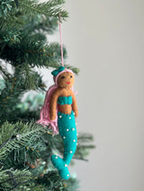 A colorful Pride Mermaid felt ornament hangs on a lush green Christmas tree. The mermaid features a smiling face with brown skin and vibrant pink wool felt hair topped with a green bow. It wears a bold turquoise top and a matching turquoise tail adorned with white dots. A pink cotton string is attached for hanging.
