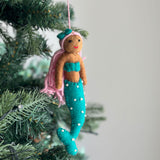 A colorful Pride Mermaid felt ornament hangs on a lush green Christmas tree. The mermaid features a smiling face with brown skin and vibrant pink wool felt hair topped with a green bow. It wears a bold turquoise top and a matching turquoise tail adorned with white dots. A pink cotton string is attached for hanging.