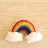 A colorful felt rainbow Christmas ornament rests on a light wood background. This handmade needle felted ornament features 3D concentric arcs of red, orange, yellow, green, and purple. Fluffy white clouds are at each end of the rainbow, with a white cotton string attached at the top for hanging.