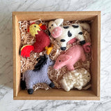 A needle felted rooster ornament is nestled in a simple brown cardboard box filled with light brown paper shred, set against a light marble backdrop. The handmade wool felt rooster ornament features a bright yellow head, a red body, and tail feathers detailed in red and black, showcasing a charming handmade aesthetic.
