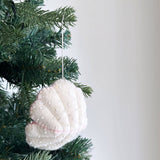 A white handmade wool felt scallop ornament hangs gracefully from a festive Christmas tree branch. This unique felt ornament features delicate rows of tiny white beads along its scalloped edges, subtly outlined with fine pink stitching. A white cotton string secures the charming ornament to the green pine needles, enhancing its rustic elegance.
