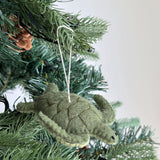 A textured light green wool felt sea turtle ornament hangs gracefully on a Christmas tree. Darker green stitching outlines its shell segments, and a white cotton string is attached for easy hanging, adding rustic charm to the holiday decor.