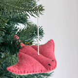 A pink wool felt stingray ornament hangs on a lush green Christmas tree, displayed against a bright white background. This charming sea animal felt ornament features a textured surface, meticulously stitched edges, small black stitched eyes, and a white cotton string for hanging.