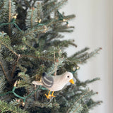 A fluffy felt seagull ornament hangs on a Christmas tree adorned with warm lights. The charming seagull has a pale body with grey wings and tail accents, complemented by a bright yellow beak and feet. A natural jute string is attached for hanging, creating a cozy and festive holiday scene.