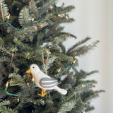 A felt seagull ornament hangs from a lit Christmas tree branch. The unique Christmas ornament features a fluffy white body, gray wings with darker gray stripes, black eyes, and a yellow beak and feet. A jute string allows the handcrafted felt ornament to gracefully hang amidst the evergreen needles and soft lights.