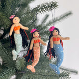 Three vibrant Silk Sea Mermaid felt ornaments are shown hanging joyfully on a green Christmas tree branch. Each handmade felt ornament features flowing black yarn hair, stitched orange and yellow seashell tops, and tails in light blue, peach, and light green adorned with white pearls. A clear string attaches them to the branch, set against a soft white background.