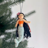 A Silk Sea felt mermaid ornament hangs gracefully on a Christmas tree. This unique handmade ornament features a soft tan face with a stitched red smile and flowing black yarn hair. It has an orange seashell top and a light blue-green tail adorned with white pearl-like embellishments. A black cotton string is attached at the top, ready for display.