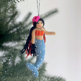 A handmade mermaid felt ornament hangs against the soft blur of a Christmas tree. This joyful felt ornament features black yarn hair, a stitched seashell top, and a blue tail adorned with pearl-like details, enhancing its handcrafted charm.