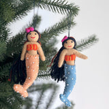 Two vibrant Silk Sea Mermaid felt ornaments are shown hanging joyfully on a green Christmas tree branch. Each handmade felt ornament features flowing black yarn hair, stitched orange and yellow seashell tops, and tails in light blue, peach, and light green adorned with white pearls. A clear string attaches them to the branch, set against a soft white background.