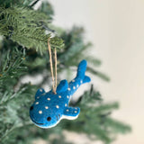 A playful, close-up view of a smiley whale shark felt ornament hangs from a green Christmas tree branch against a soft white background. This handcrafted light blue felt whale shark is adorned with white polka dots, small black eyes, and a stitched black smile, creating a friendly and cheerful appearance.