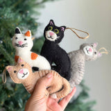 A female hand holds four charming wool felt stitched cat ornaments against a soft-focus Christmas tree background. One light brown cat ornament lies horizontally in the palm, while a white and orange, a black, and a grey cat ornament are stacked vertically. Each unique Christmas tree ornament features delicate stitched eyes, nose, and whiskers, suspended by a natural jute string.