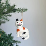 A handcrafted felt cat ornament, with its distinct white, black, and orange spots, hangs charmingly from a green Christmas tree branch. This wool felt cat ornament features a pink nose and delicate hand-stitched black whiskers, offering a warm and festive accent.