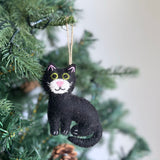 A cozy Wool Felt Stitched Cat Ornament, featuring a black body, upright pink ears, white whiskers, and white paws, hangs from a jute string on a Christmas tree. The cat's white face has a pink nose and yellow eyes with green pupils. Subtle lights and pinecones adorn the festive tree.