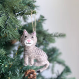 A stitched cat felt ornament hangs on a vibrant green Christmas tree, creating a cozy and festive atmosphere. This gray wool felt Christmas ornament features intricate hand-stitched details, a pink nose, black eyes, and gray stripes. A jute string loop is attached for hanging. The blurred Christmas tree, adorned with small lights, adds to the warm, inviting feel.
