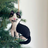 A wool felt stitched cat ornament hangs from a jute string against the cheerful backdrop of a Christmas tree. This charming black and white cat ornament features pointy ears, a tiny pink nose, and embroidered whiskers, adding a warm, playful touch. The handcrafted felt ornament evokes homemade charm and holiday joy.