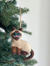 A Wool Felt Stitched Cat Ornament hangs from a green Christmas tree, set against a clean, light background. This cream-colored cat has dark brown ears, a facial mask, legs, and tail. It features hand-stitched bright blue eyes, white whiskers, and a pink nose.