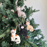 Various Wool Felt Stitched Cat Ornaments adorn a green Christmas tree, creating a cozy, festive display. Each unique handmade cat ornament, including gray striped, orange striped, black, and white with orange spots, features intricate hand-stitched details and expressions. They are suspended by jute strings among pine cones.