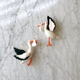 Handcrafted felt stork ornaments are displayed on a mottled white marble surface. Each white stork features an orange beak and legs, detailed black and white felt wings, and holds a small white bundle tied with either a red or white string. A hemp string is attached, ready for hanging.