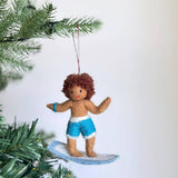 A handmade felt surfer boy ornament hangs from a Christmas tree branch by a brown cotton string. This charming felt ornament features a surfer boy with brown curly hair, brown skin, a red smile, and black eyes. He wears a blue arm band, blue and white shorts, and stands on a blue and white surfboard.