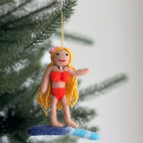 A playful surfer girl felt ornament hangs by a gold string from a green Christmas tree branch. This handcrafted ornament features a tan-skinned figure with golden hair, a red smile, a pink flower on her head, and a pearl choker necklace. She wears a red bikini and stands confidently on a blue and white surfboard, bringing a sense of summer fun to the holiday setting.
